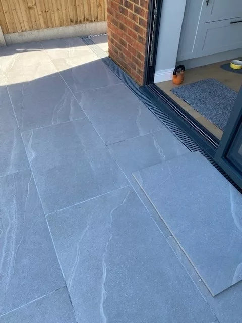 Large Ashmore Grey Porcelain Slabs create a stylish patio leading into a modern kitchen through open glass doors. A drain separates the patio from the interior, where part of a rug and kitchen appliances are visible inside.