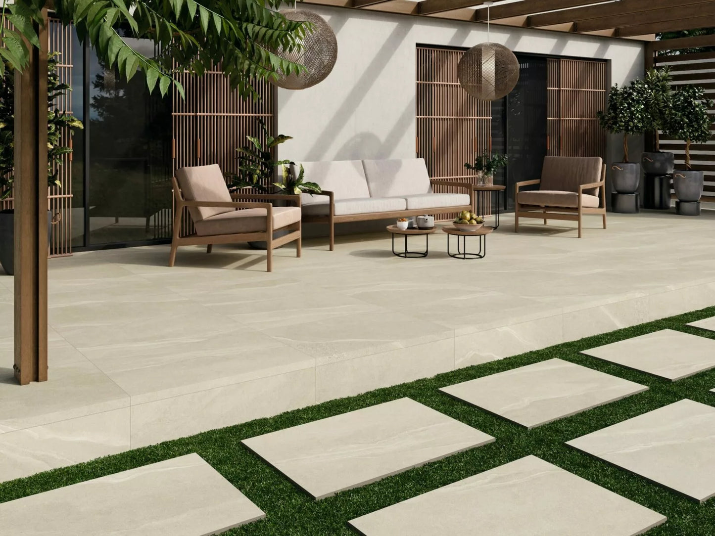 Modern outdoor patio with Ashmore Ivory Porcelain Paving Slabs, wooden furniture with beige cushions, round coffee tables, hanging lanterns, and lush green plants; stone pavers lead across a grassy area in the foreground.