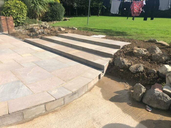 Partially completed outdoor stone steps with Autumn Brown Sandstone Paving leading to a grassy yard; construction materials and rocks are visible on the right, and laundry hangs on a clothesline in the background.