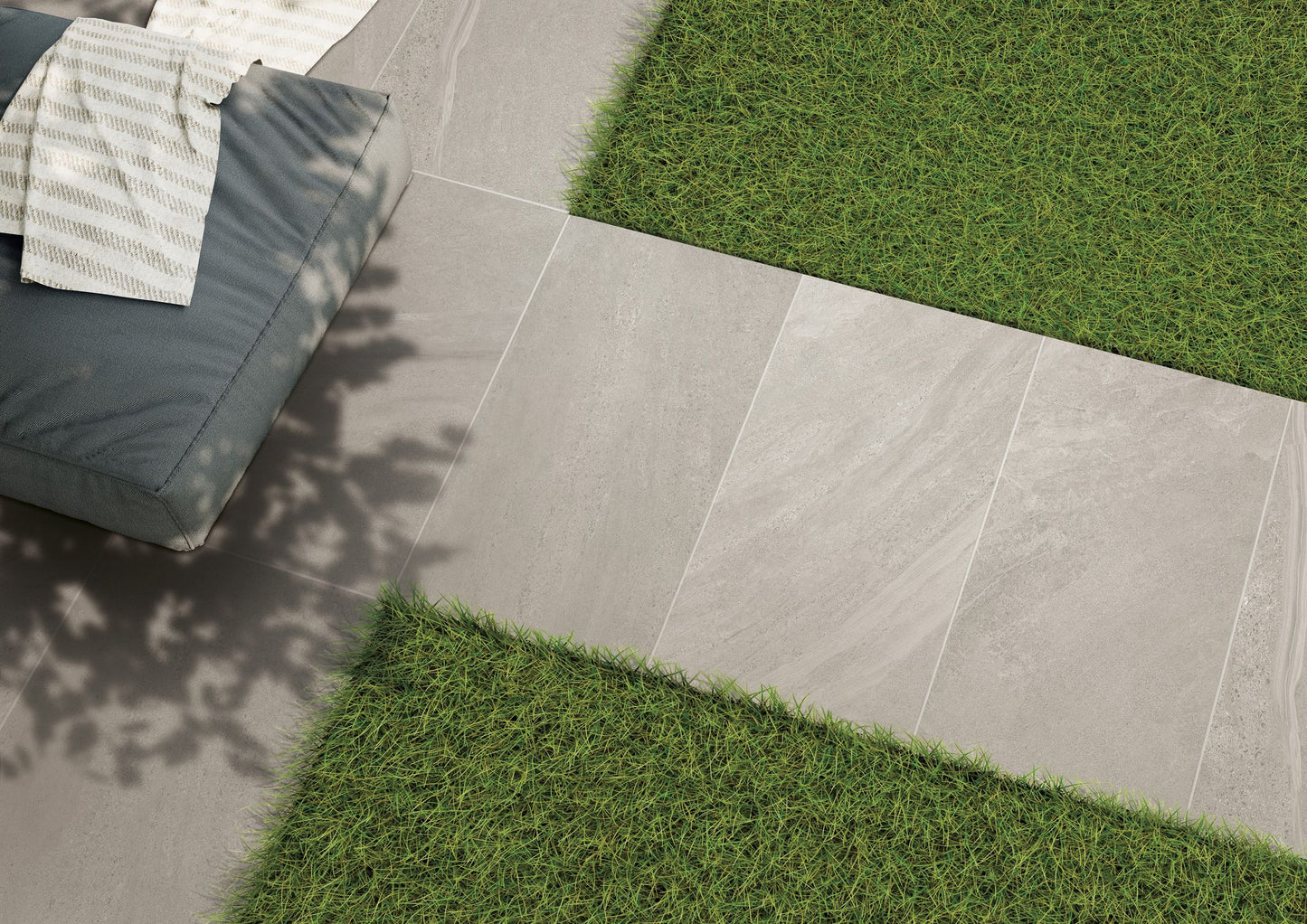 Gray rectangular slip resistant tiles form a path bordered by green grass. Part of a blue cushioned seat with a striped white towel is visible in the upper left corner, with some soft shadow from nearby foliage.