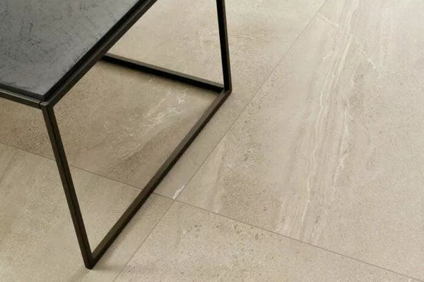 A close-up view of beige travertine-look outdoor porcelain tiles with subtle natural patterns, next to the corner of a black metal table with a dark tabletop.