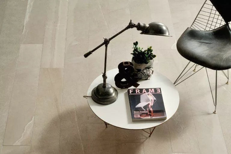 A round white table with a vintage lamp, potted plant, decorative ampersand, and "FRAME" magazine sits next to a black wire chair on light-colored slip resistant pavers.
