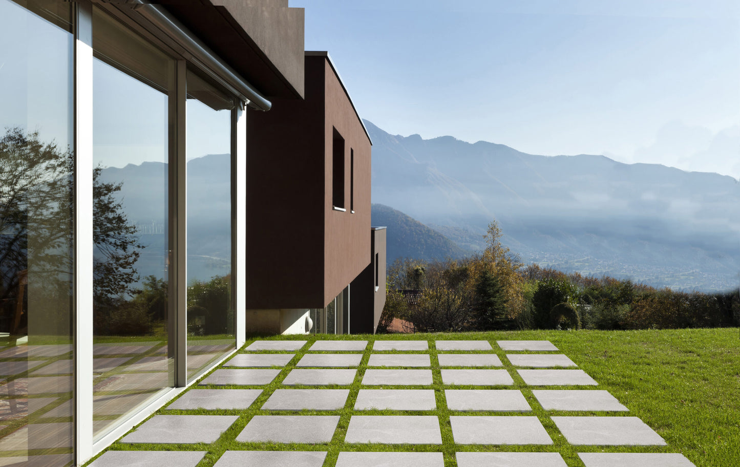 Modern house with large glass windows and brown exterior, overlooking a garden with Brampton Perla Porcelain tiles and green grass. In the background, misty mountains rise beneath a clear sky, enhancing the home's stylish use of outdoor porcelain tiles.