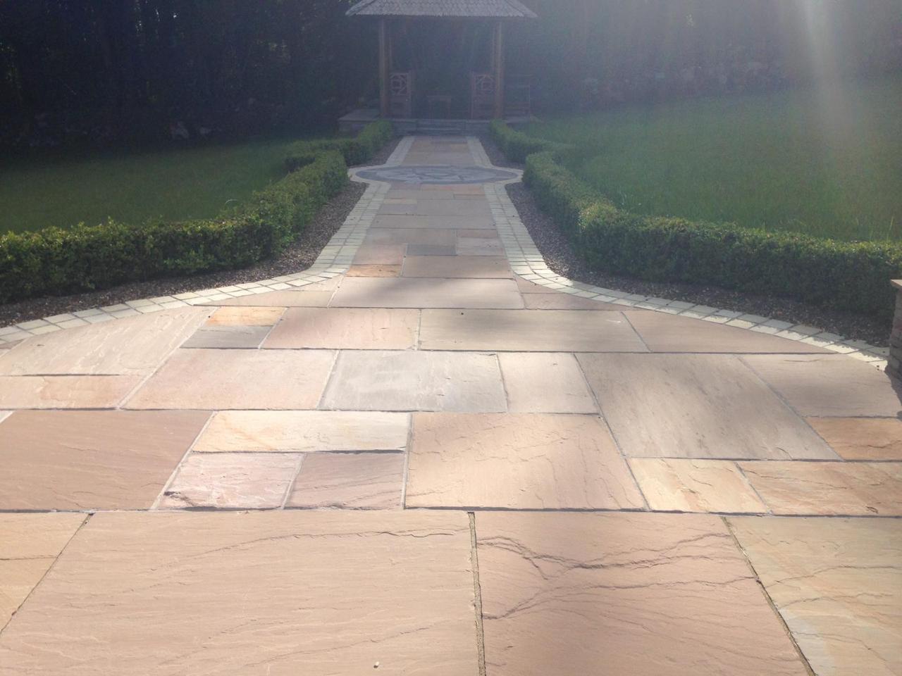 A stone pathway with large buff sandstone paving tiles leads through a manicured lawn bordered by hedges toward a wooden gazebo in the background. Sunlight creates soft shadows on the path.