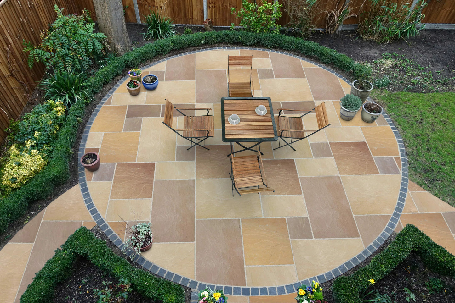 A circular patio with Buff Sandstone Paving tiles, bordered by dark stone, features a table with four wooden chairs. Potted plants and trimmed hedges surround the patio, with a lawn and garden beds nearby.