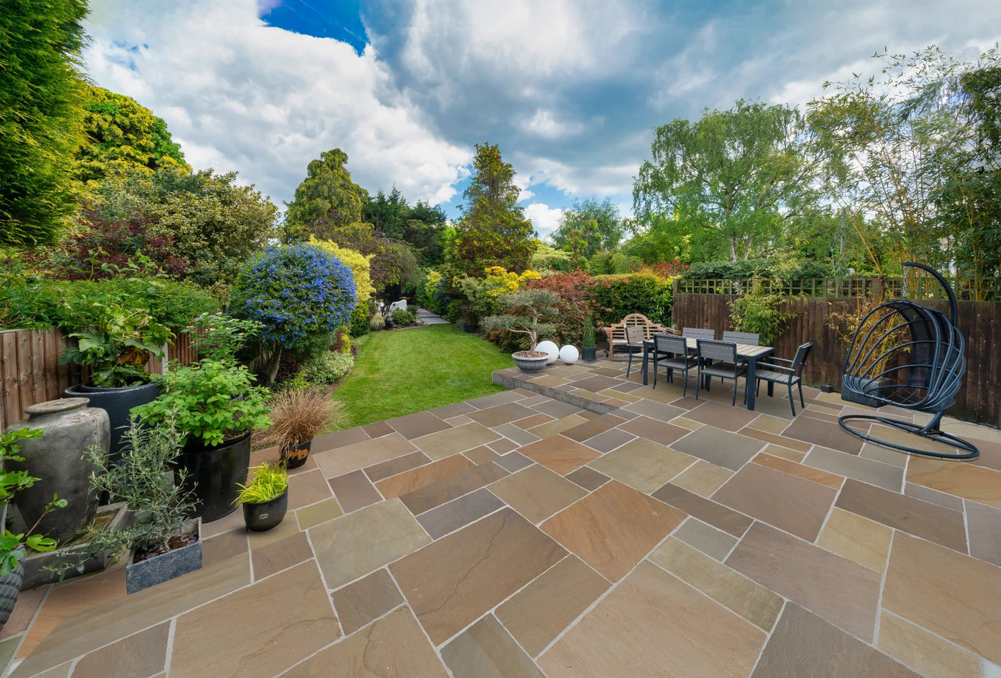 Spacious backyard with a stone patio featuring Buff Sandstone Paving, an outdoor dining table, hanging chair, potted plants, and a lush green lawn bordered by trees, shrubs, and colorful flowers under a partly cloudy sky.