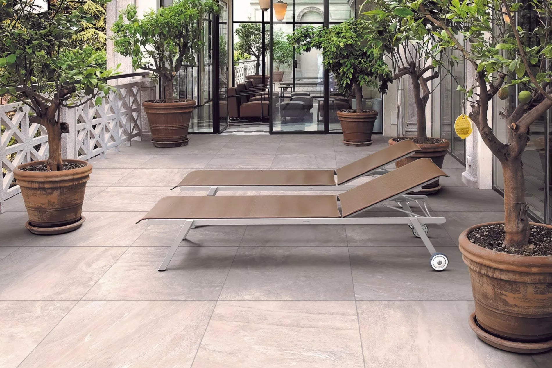 Two brown lounge chairs sit on a tiled patio surrounded by large potted trees, with glass doors in the background leading to an indoor seating area. The space is bright and inviting.