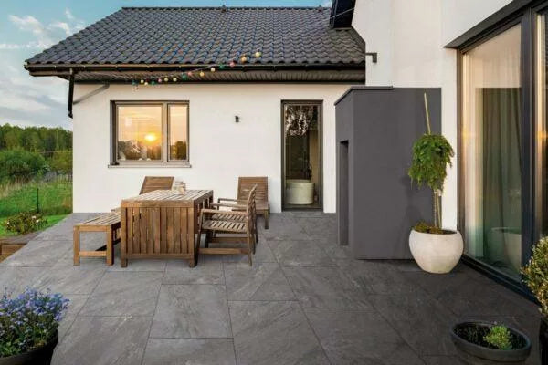 A modern outdoor patio with grey porcelain paving, wooden dining table and chairs, potted plants, and string lights; the house has white walls and large windows, with a sunset reflecting in the window.