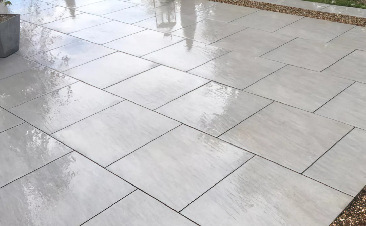 Wet, light gray Fraser Grigio Porcelain Paving Slabs are neatly arranged outdoors in a staggered pattern, reflecting light. Gravel borders the edge, and some plants and a planter are partially visible.