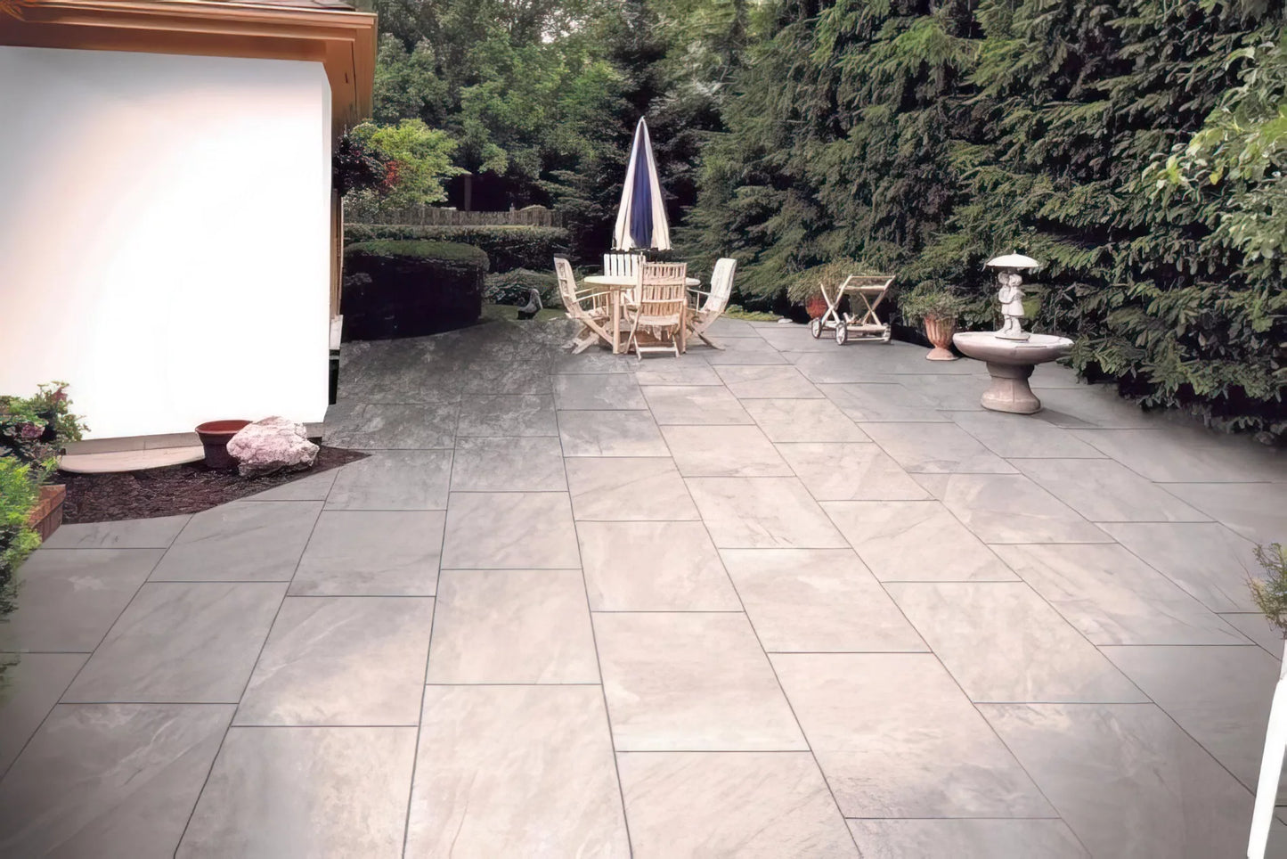 Spacious patio with Fraser Grigio Porcelain Paving Slabs, an outdoor wooden dining set with umbrella, small table, fountain, and lush greenery with tall trees surrounding the area.
