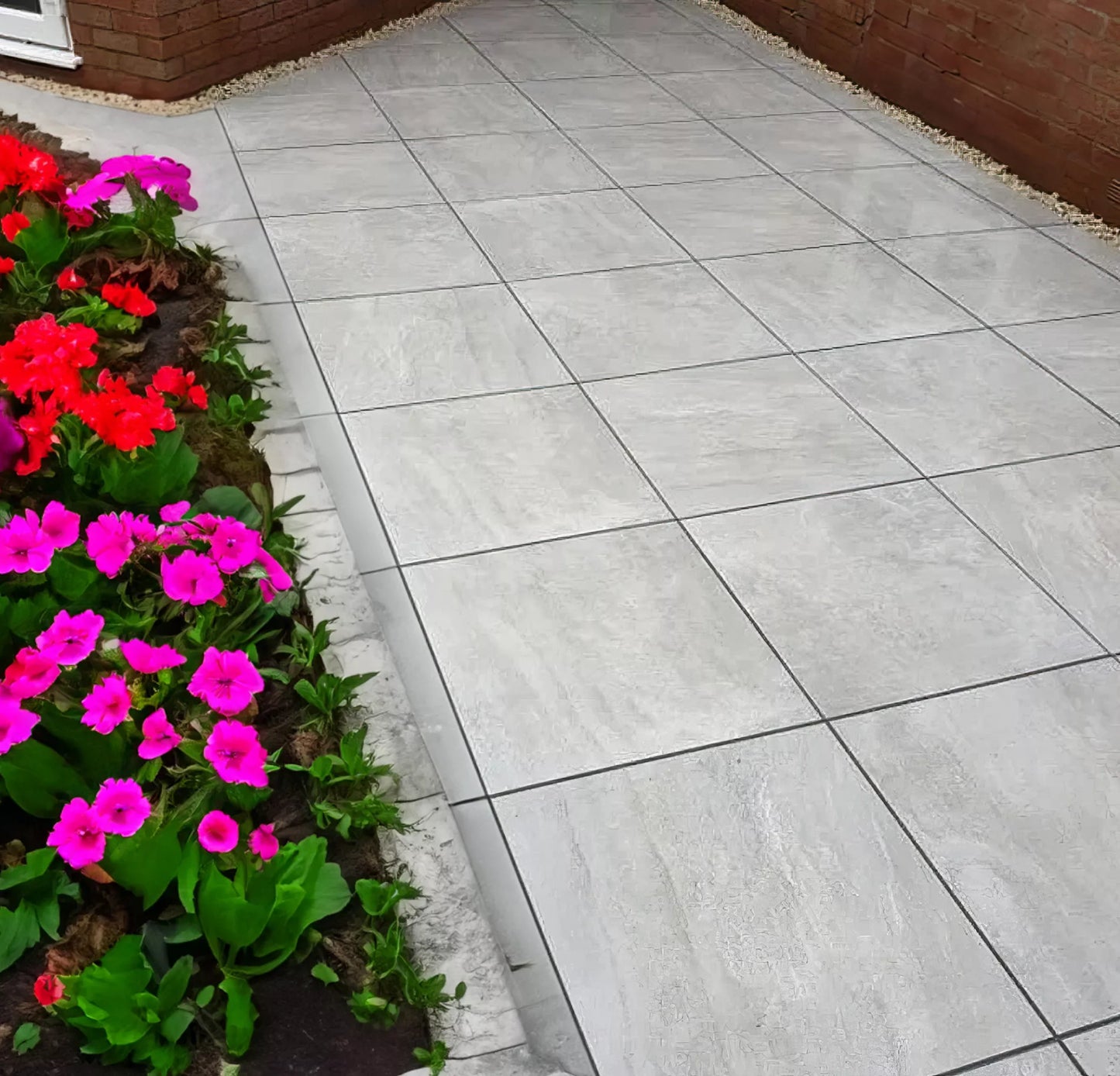 A garden with vibrant pink and red flowers borders a clean, light gray patio featuring Fraser Grigio Porcelain Paving Slabs next to a brick wall.
