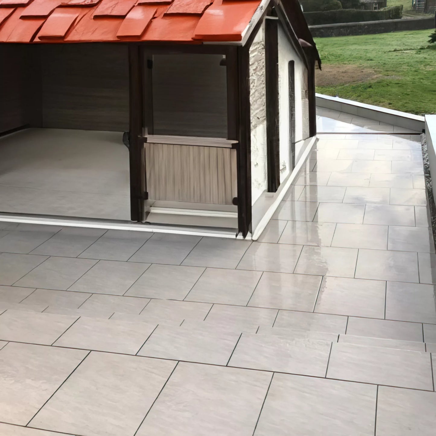 A large patio with light gray Fraser Grigio Porcelain Paving Slabs extends beside a small structure with a red tiled roof and partially open walls, overlooking a grassy area and distant shrubs.