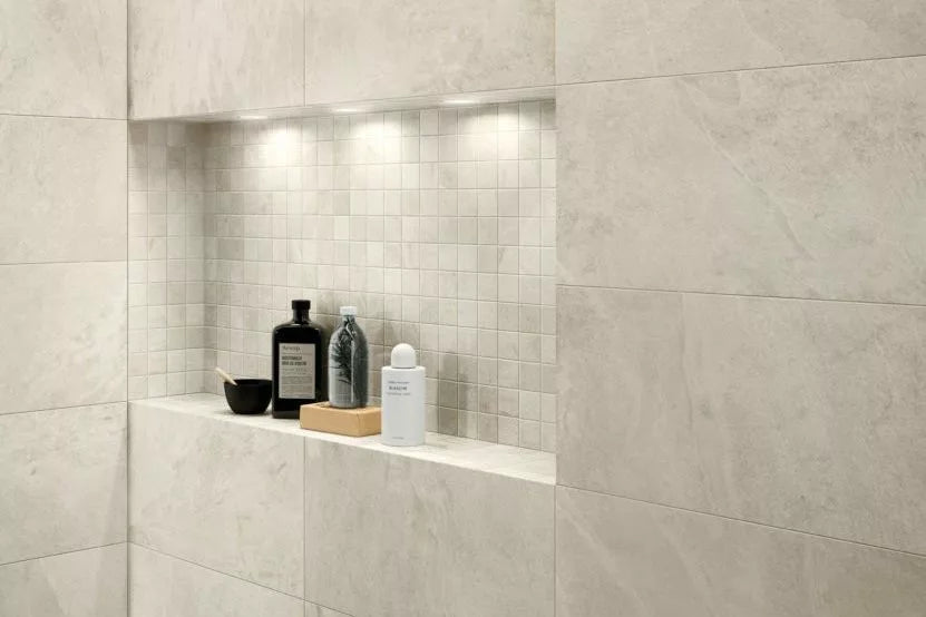 A recessed shower shelf with beige tiles, reminiscent of outdoor pavers, contains black and white bottles, a clear bottle, a wooden brush, and a small black bowl. The shelf is illuminated by three built-in lights.