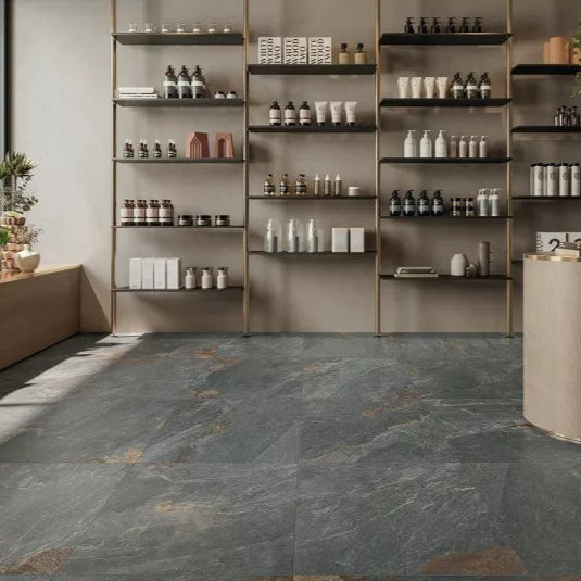 Modern store interior with stone tile flooring resembling porcelain paving, wooden shelves on the back wall displaying neatly arranged bottles and jars, and a curved checkout counter on the right. Natural light enters from the left.