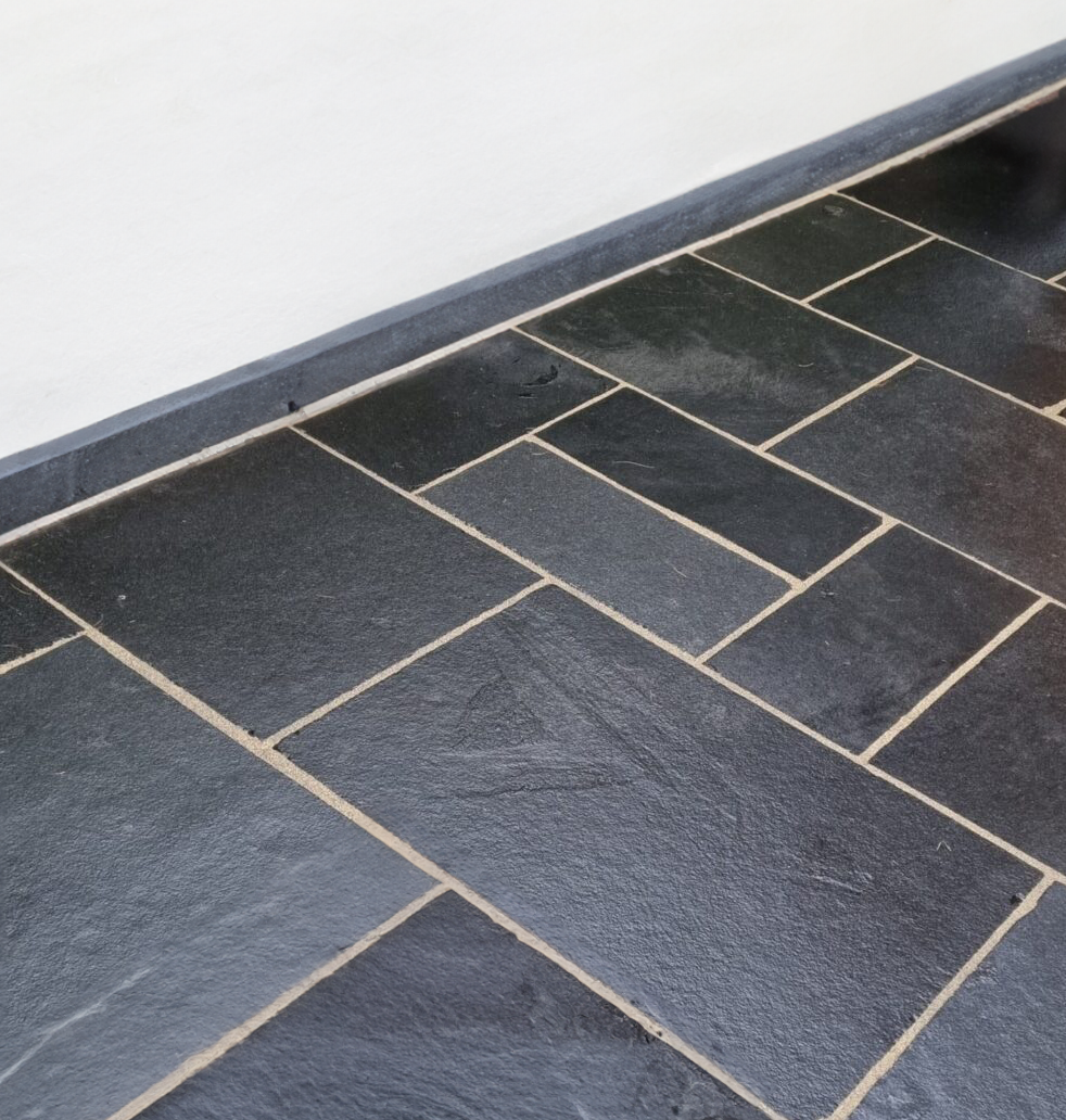 Dark slate paving tiles with light-colored grout arranged in a geometric, staggered pattern along the edge of a white wall and dark baseboard evoke the sleek look of Kadappa Black Limestone Paving.