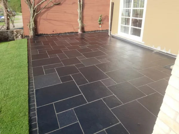 A patio with Kadappa Black Limestone Paving tiles arranged in a geometric pattern beside an artificial grass lawn, bordered by two beige walls and a glass door. Two leafless trees stand near the corner.