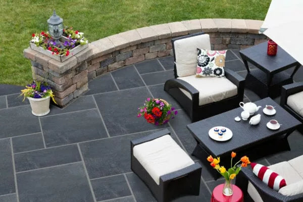 A modern patio with black wicker furniture, cream cushions, a black table set for tea and desserts, fresh flowers in vases, and a curved stone wall with potted plants; garden stepping stones lead to green grass beyond the wall.