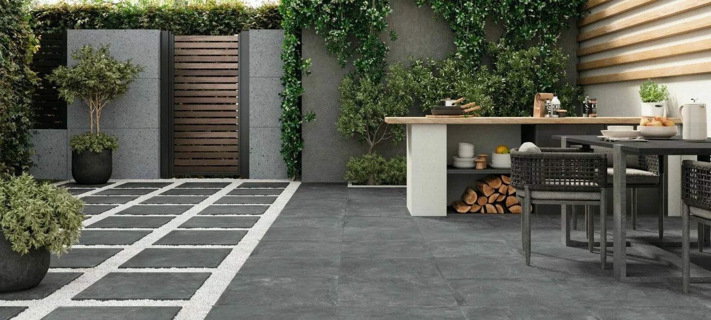 Modern outdoor patio with stylish Palmer Graphite Porcelain Paving Slabs, a dining table with woven chairs, potted plants, a small outdoor kitchen with wood storage, and lush greenery along the fence and walls.