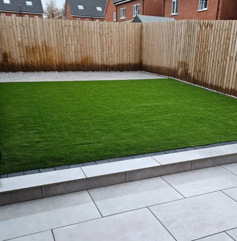 A neatly landscaped backyard with a rectangular patch of green grass bordered by Palmer Grey Porcelain Paving Slabs and a wooden fence. Brick houses are visible in the background, adding charm to this inviting outdoor space.