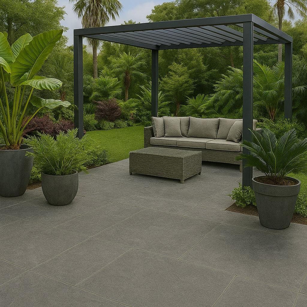 A modern outdoor patio with Palmer Graphite Porcelain Paving Slabs, a gray pergola, wicker sofa with cushions, matching coffee table, and several potted tropical plants. Lush green trees and grass surround the area.