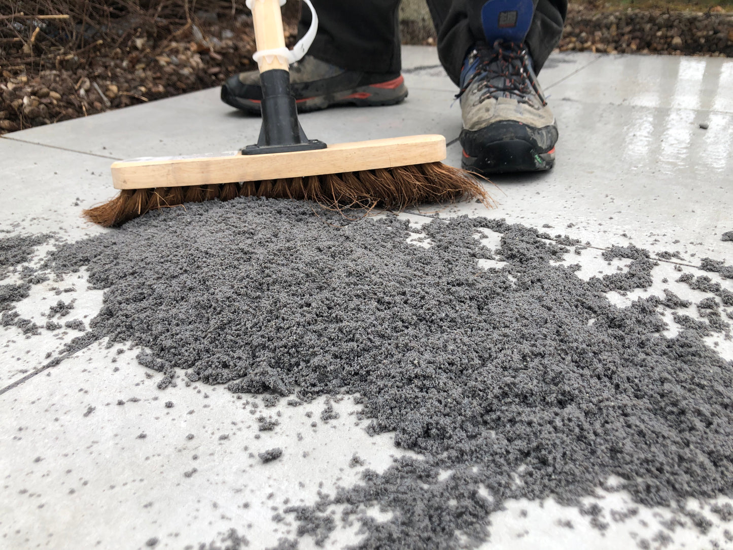 A person wearing outdoor shoes sweeps gray sand or fine gravel, possibly as a brush-in jointing solution, across large stone tiles with a broom on a patio or sidewalk.