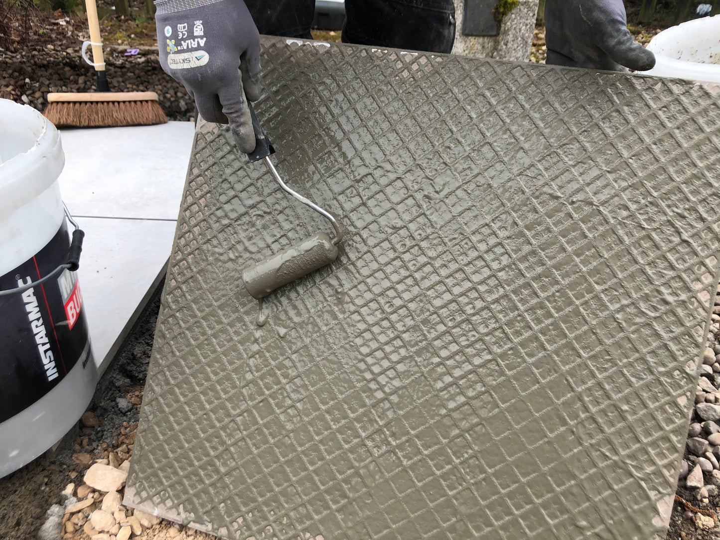 A person wearing gloves uses a small paint roller to apply ProPave Primer over a textured outdoor tile as part of an external tiling system, with a white bucket and broom visible in the background.