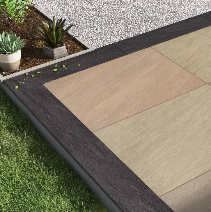 A modern patio with large beige and tan Raj Green Porcelain Paving Slabs bordered by dark tiles, next to green grass and a gravel area with potted succulent plants.
