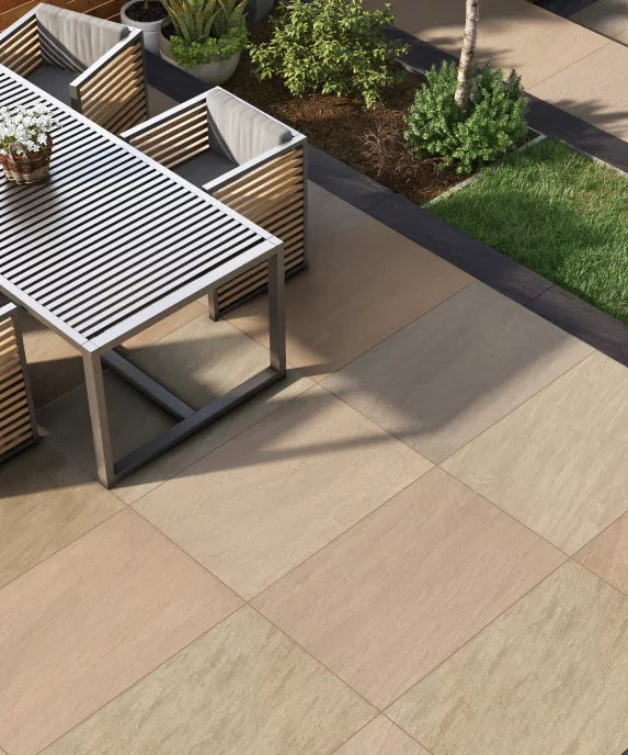 Modern outdoor patio with large beige and tan Raj Green Porcelain Paving Slabs, a slatted metal table, striped cushioned chairs, potted plants, and landscaped greenery in the background.