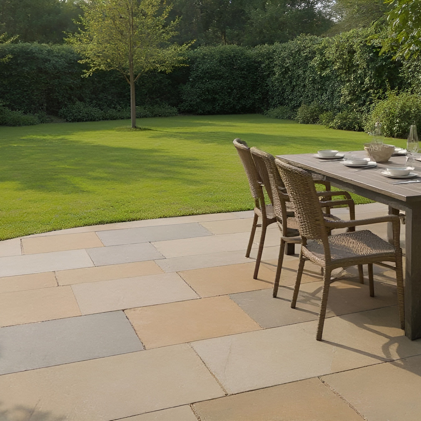 A patio with a stone floor of Raj Green Sandstone Paving features a wooden dining table and wicker chairs set for a meal, overlooking a neatly trimmed lawn with trees and dense green hedges in the background.