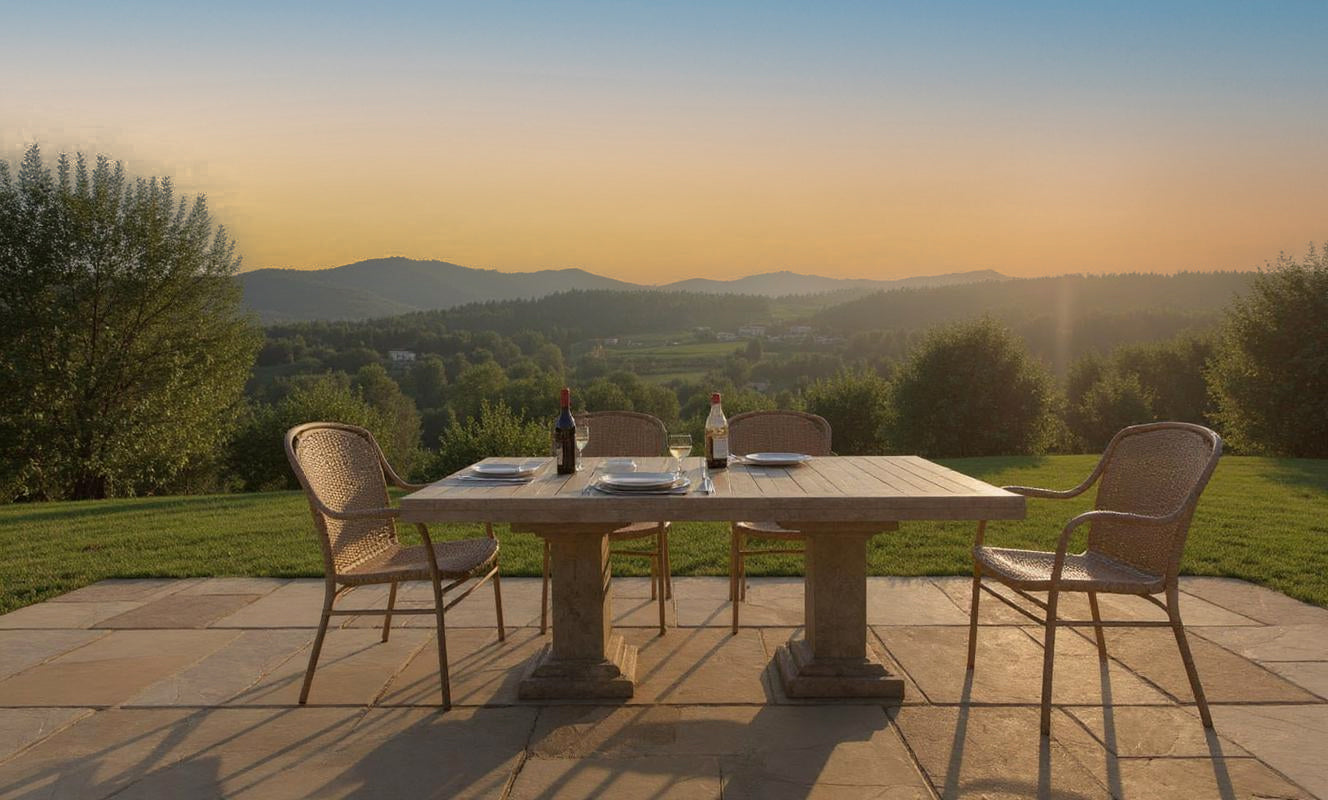 A wooden outdoor dining table with four chairs is set with plates, glasses, and two wine bottles on Raj Green Sandstone Paving, overlooking a scenic landscape of hills and trees at sunset.