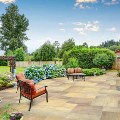 A spacious backyard patio with tan Raj Green Sandstone Paving tiles, cushioned wrought iron chairs, flower beds of blooming hydrangeas, green shrubs, and a lawn bordered by trees under a partly cloudy blue sky.