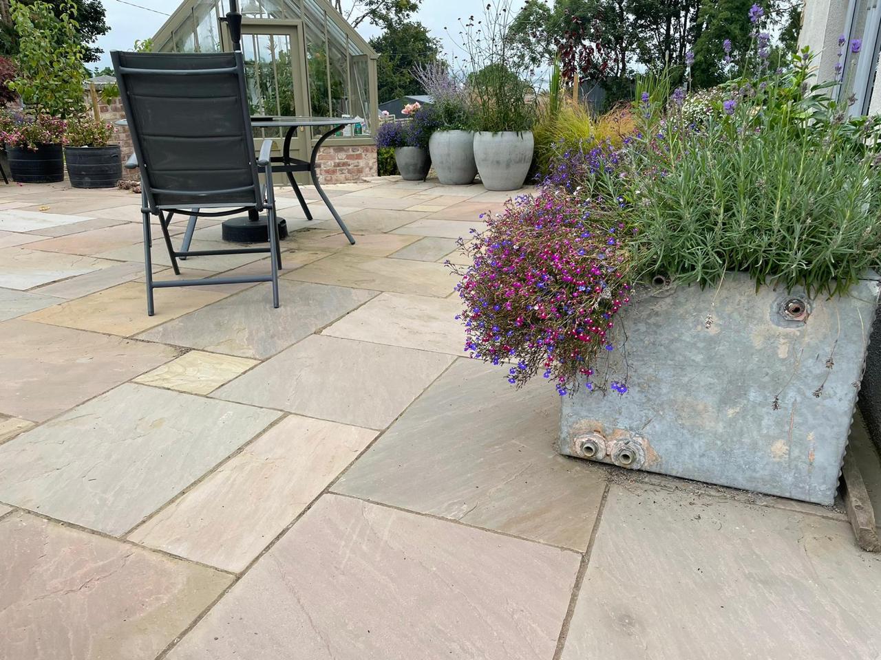 A stone patio with Raj Green Sandstone Paving, potted plants and flowers, a metal table with chairs, and a greenhouse in the background. The patio is surrounded by garden greenery and large planters.