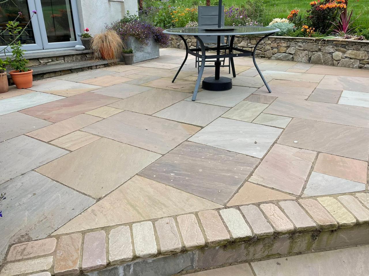 A stone patio with Raj Green Sandstone Paving, a round metal table and chairs, potted plants, and a raised garden bed with flowers and shrubs along a stone wall. Steps lead up to a glass door.