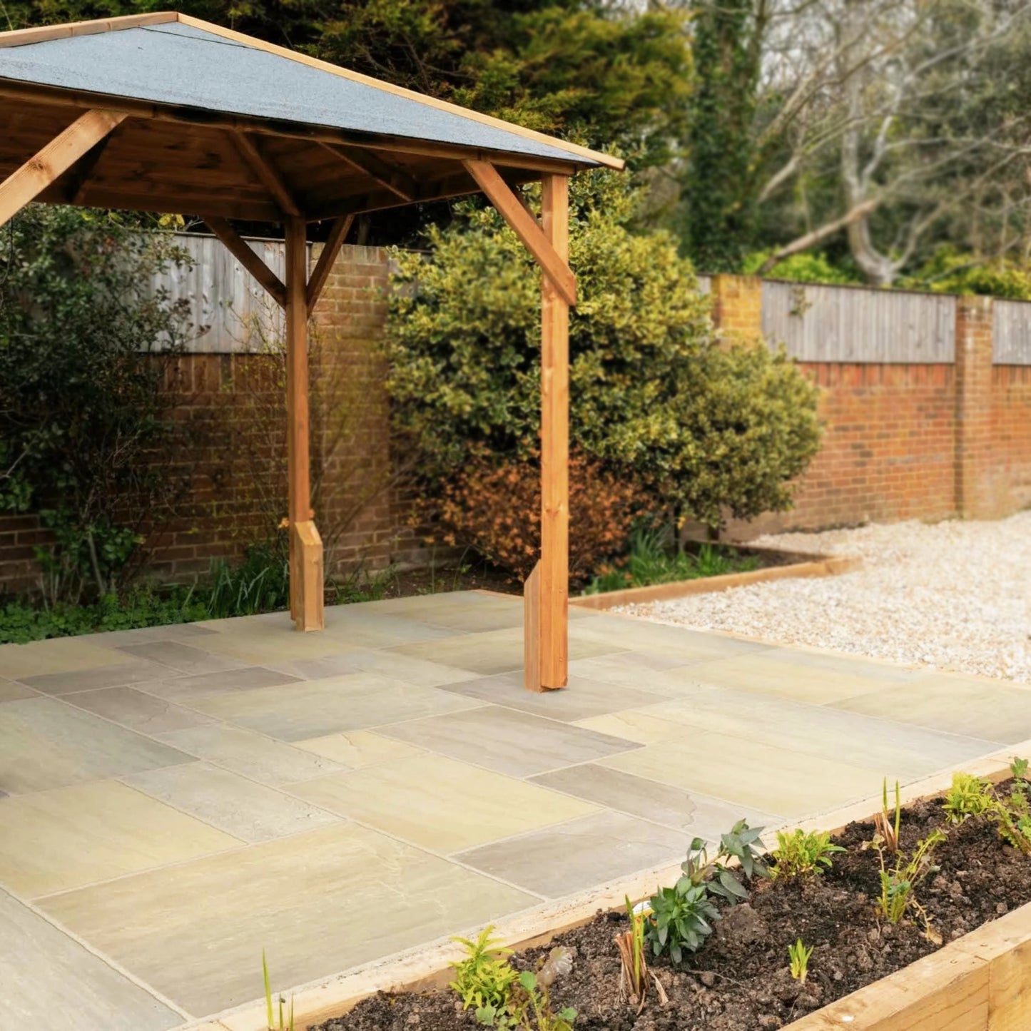 A wooden gazebo with a slate roof stands on a stone patio in a garden, enhanced by Raj Green Sandstone Paving and bordered by a low brick wall, gravel area, green shrubs, and a small planted flowerbed in the foreground.