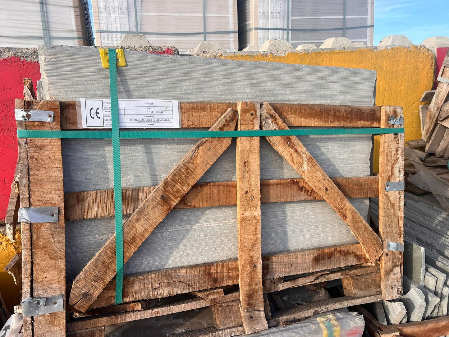 A stack of large, rectangular gray paving tiles secured with green straps and a wooden frame, placed outdoors among other building materials and colorful concrete barriers.