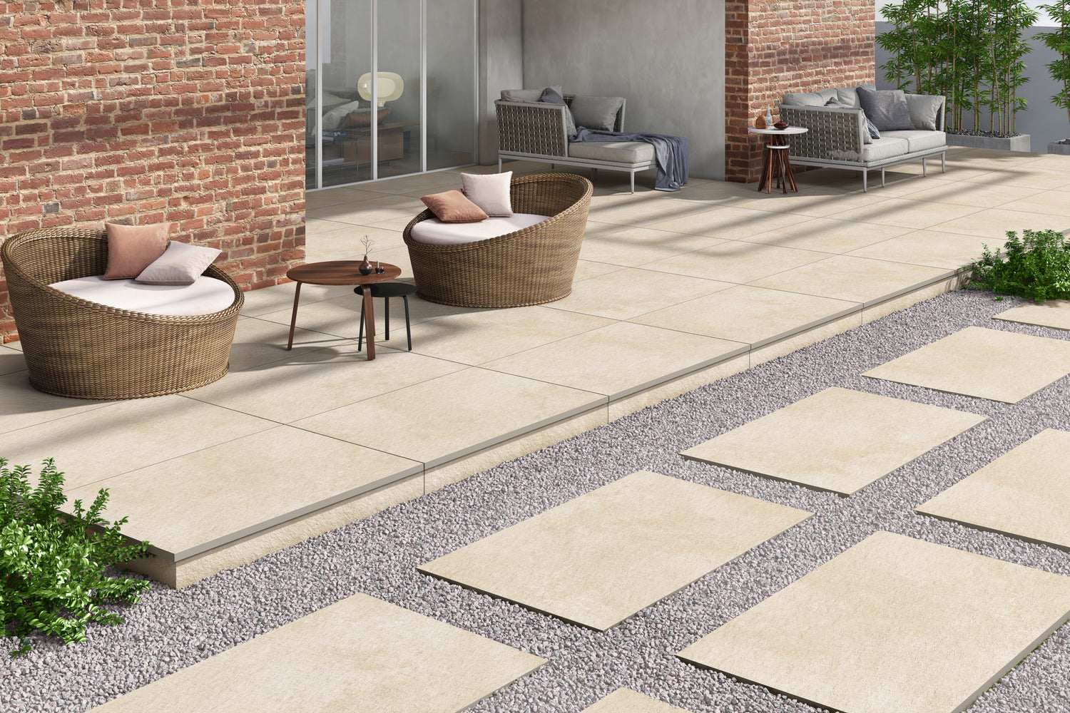 Modern outdoor patio with beige porcelain paving slabs, round wicker chairs with white cushions and pillows, a small wooden table, sofas, and stepping stones set in gravel, surrounded by greenery and bordered by a brick wall.