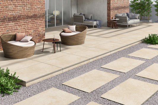 Modern outdoor patio with beige porcelain paving slabs, round wicker chairs with white cushions and pillows, a small wooden table, sofas, and stepping stones set in gravel, surrounded by greenery and bordered by a brick wall.