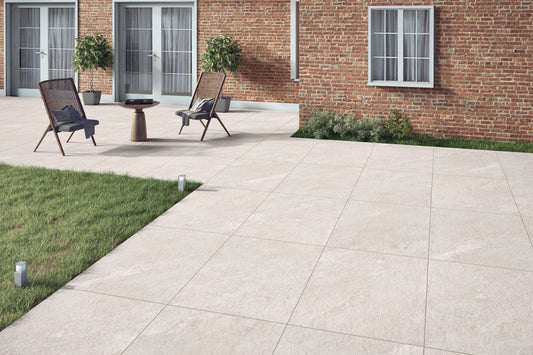 A modern patio with Antigua White Porcelain Paving Slabs next to a brick building, two chairs and a small table, potted plants, grass borders, and three glass doors with white curtains.