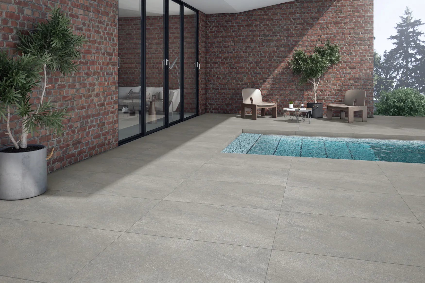 Modern outdoor patio with large Antillies Grey Porcelain Paving Slabs, a swimming pool, potted plants, and two chairs with a small table near a brick wall and glass doors leading inside. Trees are visible in the background.