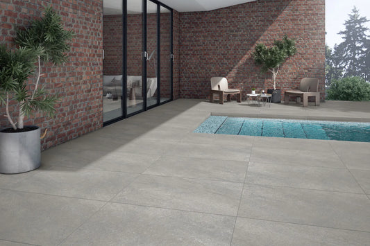 Modern outdoor patio with large Antillies Grey Porcelain Paving Slabs, a swimming pool, potted plants, and two chairs with a small table near a brick wall and glass doors leading inside. Trees are visible in the background.