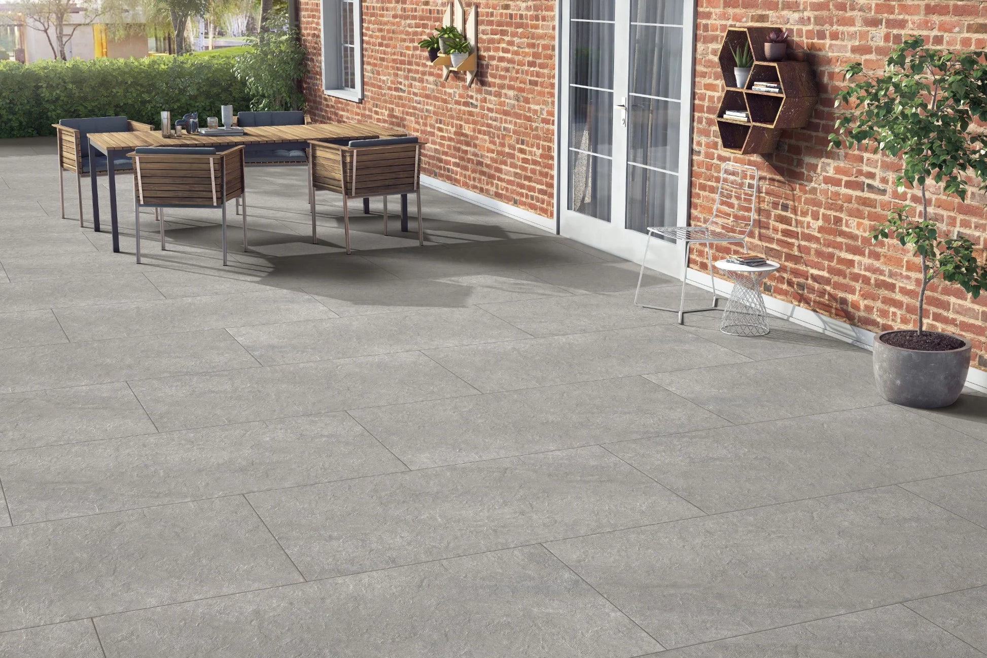 Outdoor patio with large Antillies Grey Porcelain Paving Slabs, a wooden dining table with four chairs, a plant on the table, and a brick wall with glass doors and hanging shelves. A wire chair and potted plant complete the inviting space.