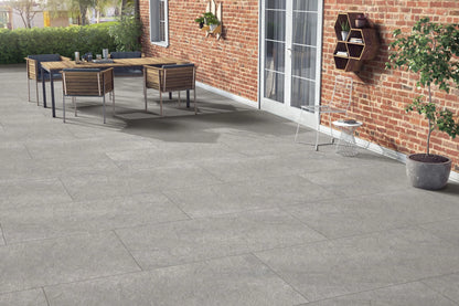 Outdoor patio with large Antillies Grey Porcelain Paving Slabs, a wooden dining table with four chairs, a plant on the table, and a brick wall with glass doors and hanging shelves. A wire chair and potted plant complete the inviting space.