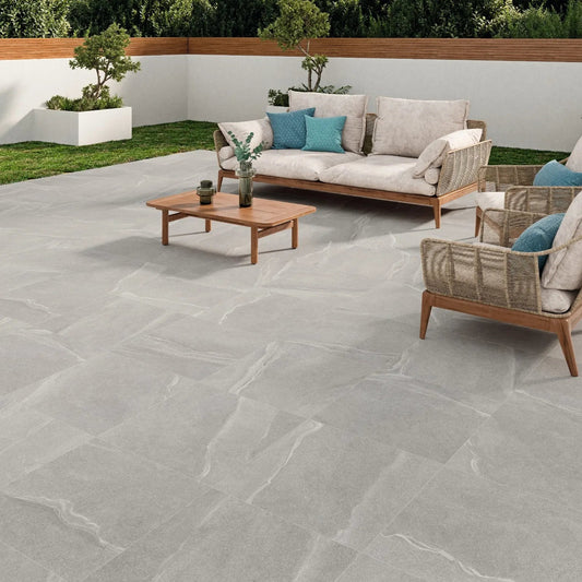 Modern outdoor patio with slip-resistant paving slabs in light gray, a wooden coffee table, a beige cushioned sofa with blue and gray pillows, an armchair, potted plants, and a white privacy wall with greenery behind it.