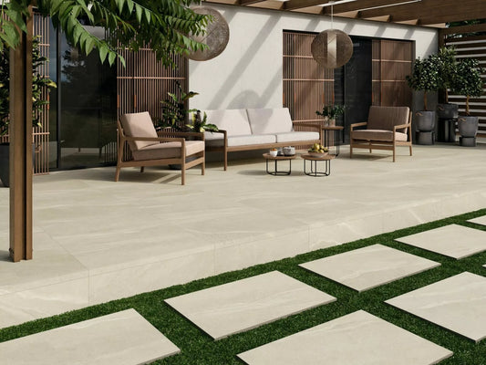 Modern outdoor patio with Ashmore Ivory Porcelain Paving Slabs, wooden furniture with beige cushions, round coffee tables, hanging lanterns, and lush green plants; stone pavers lead across a grassy area in the foreground.