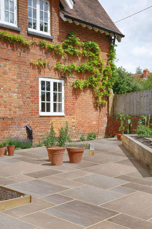 A brick house with white-framed windows, climbing greenery, a stone tile patio featuring Autumn Brown Sandstone Paving and three terracotta plant pots, a raised garden bed, and a wooden fence in the background.