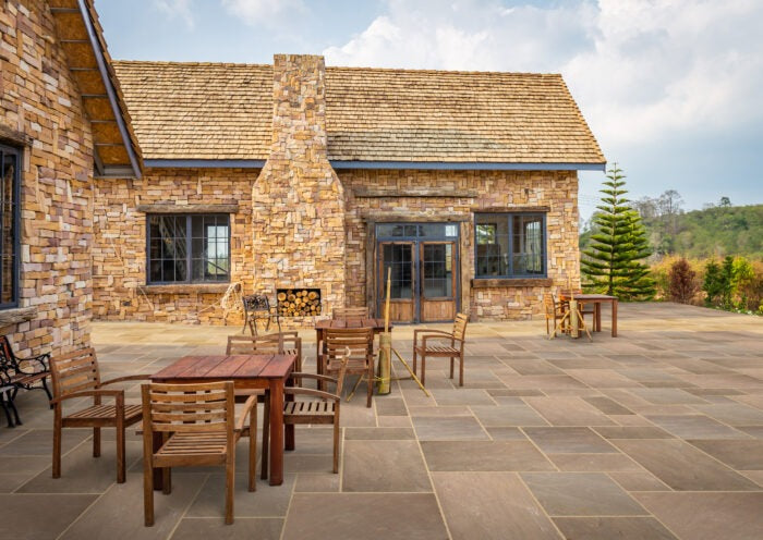 Stone patio with wooden tables and chairs outside a rustic stone house, featuring large windows and a chimney, surrounded by greenery and trees. The patio showcases Autumn Brown Sandstone Paving under a partly cloudy sky.