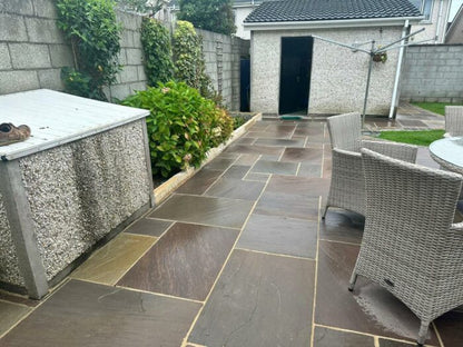 A paved backyard patio with Autumn Brown Sandstone Paving and wicker chairs around a glass table, bordered by a low wall, lush green plants, and a shed. A concrete structure sits at the left and a rotary clothesline stands in the background.