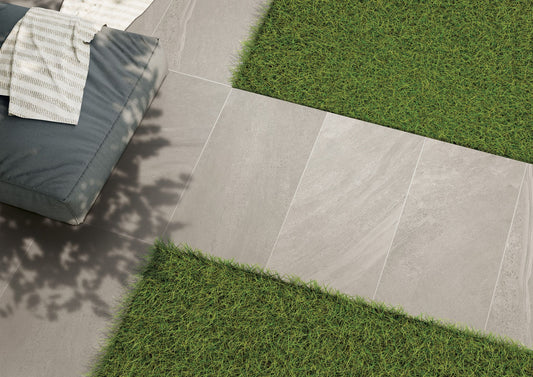 Gray rectangular slip resistant tiles form a path bordered by green grass. Part of a blue cushioned seat with a striped white towel is visible in the upper left corner, with some soft shadow from nearby foliage.