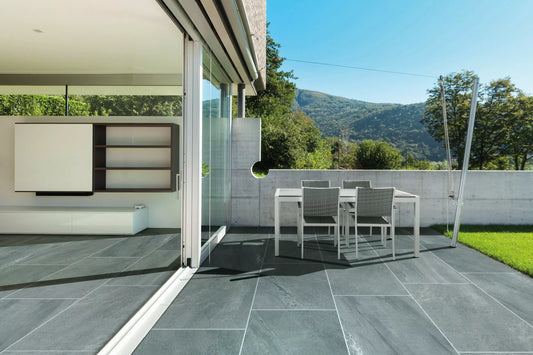 Modern patio with large gray outdoor pavers, a white dining set, and a sliding glass door opening to a minimalist living room. Green lawn and trees frame mountain views under a blue sky.