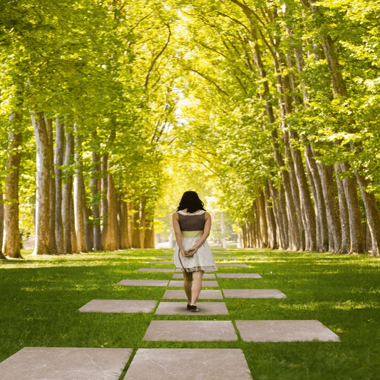 A woman walks alone on stone steps made of slip-resistant paving tiles through a lush green park, surrounded by tall trees forming a leafy canopy overhead, with sunlight filtering through the branches.